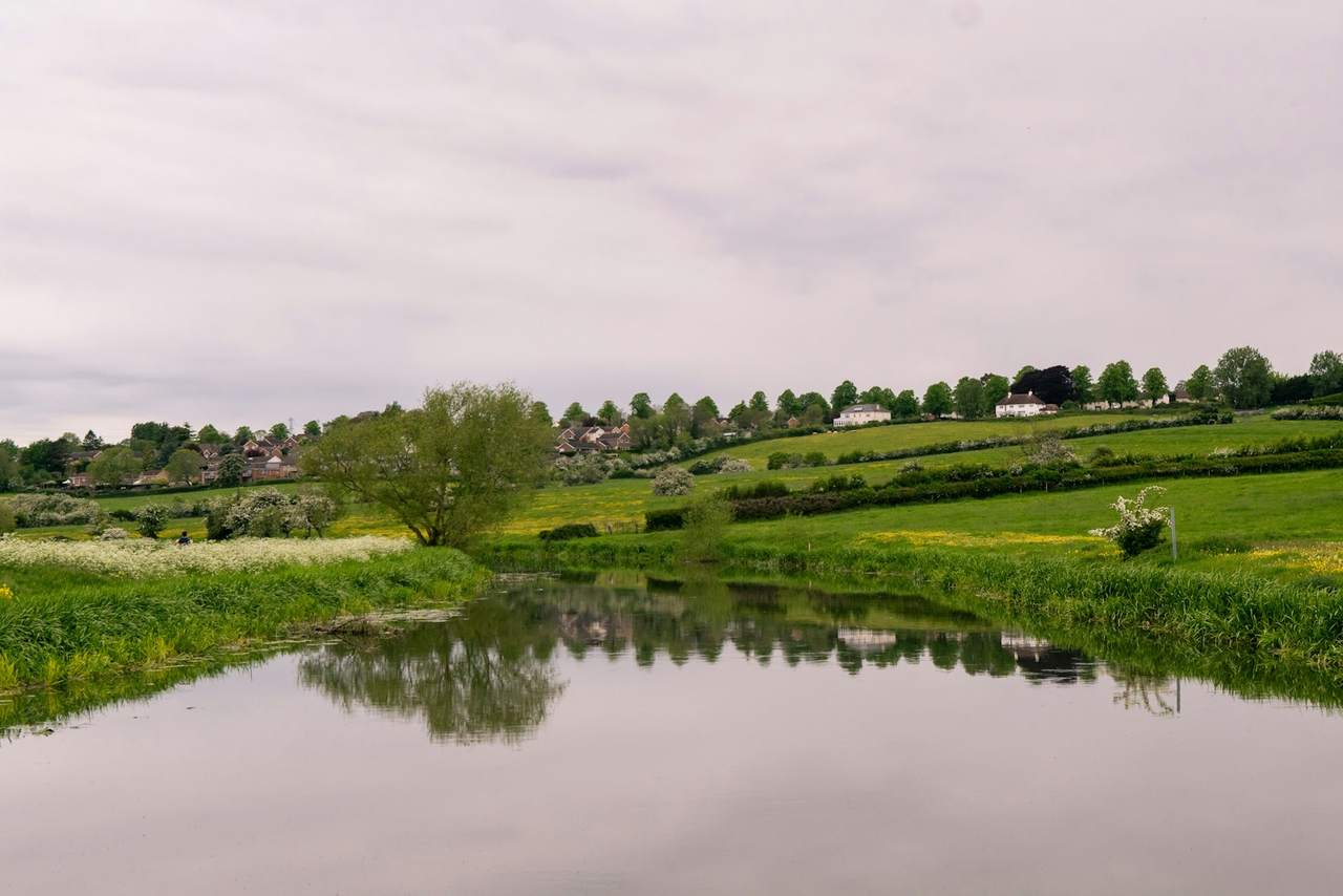 A still pond reflecting trees beside green fields under an overcast sky