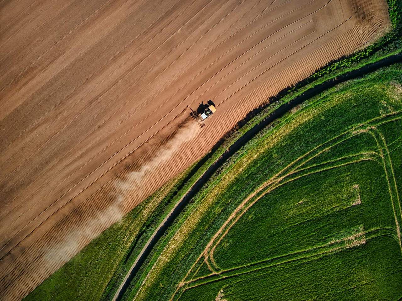 Aerial view of a tractor cultivating a bare field beside green pasture, trailing dust